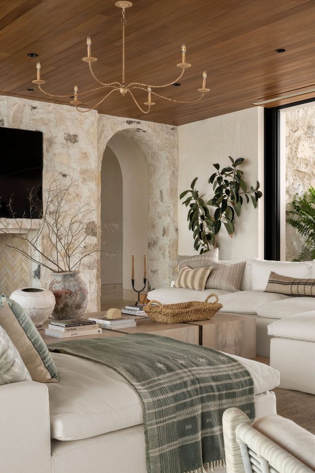 Warm, refined living room with a large cream sectional sofa and ottoman, layered neutral cushions, a green-grey throw, and a low timber coffee table. The room features a textured stone or plaster feature wall with an arched opening, a rich timber-lined ceiling, black-framed glazing, indoor plants, and an ornate suspended chandelier.