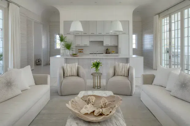 Bright, symmetrical open-plan coastal-style living room with white sofas and armchairs arranged around a low marble coffee table. The room opens directly toward a white kitchen with island cabinetry, integrated appliances, pendant lights, and a tiled or stone-look backsplash. White horizontal wall panelling, louvred shutters, extensive glazed doors and windows, pale flooring, and soft neutral furnishings create an airy, refined interior.