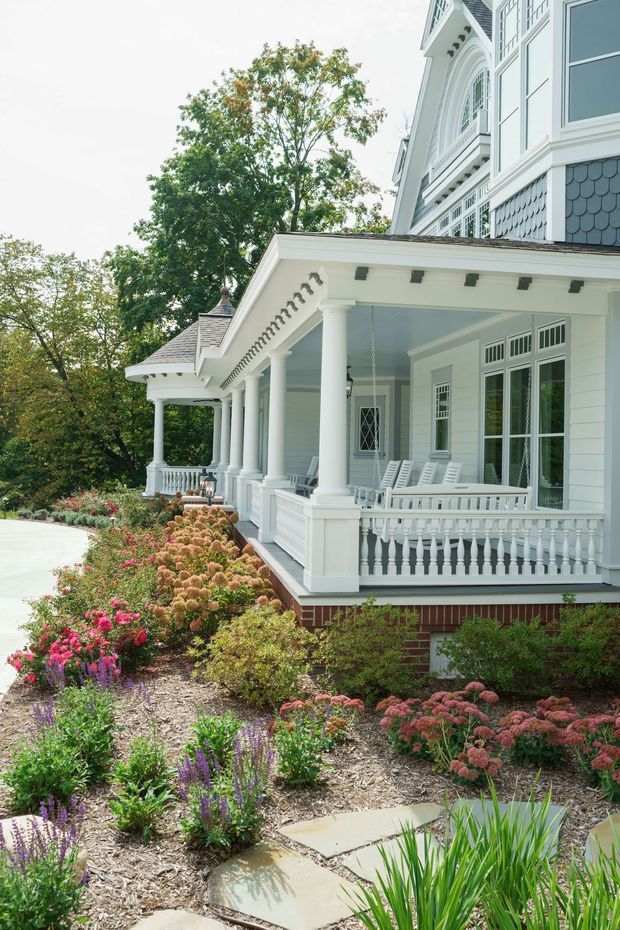 Exterior view of a large white traditional-style house with an expansive covered veranda, classical columns, decorative balustrades, ornate window trim, and shingle detailing. A landscaped garden with flowering plants and stepping-stone paths borders the veranda, while a pale driveway or walkway is visible to the left.