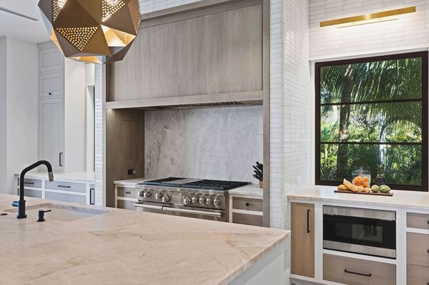 Bright, refined contemporary kitchen featuring a large pale stone island with an undermount sink and dark tapware, a professional stainless-steel range beneath a full-height stone backsplash, light wood cabinetry, white tiled walls, and integrated appliances. A dark-framed window overlooks lush greenery, while a geometric brass-and-black pendant adds a decorative focal point.