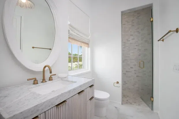 Bright, refined bathroom with a white marble vanity top, fluted white fixed cabinetry, brushed brass tapware and hardware, an oval wall mirror, and a glass-enclosed shower lined with pale mosaic tile. A toilet and window with a woven Roman shade are also visible. The space has a clean, elegant, softly traditional character.