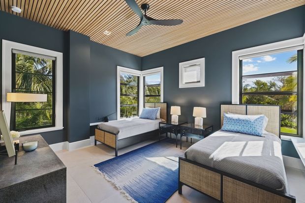 A bright, refined bedroom with two single beds, dark blue-grey walls, a slatted light timber ceiling, large windows, bedside tables, and a blue-and-white area rug. The room has a contemporary coastal feel with abundant natural light and views of tropical greenery outside.