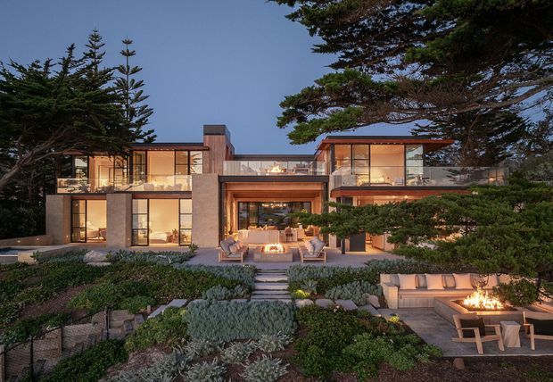A sophisticated multi-level contemporary residence photographed at dusk, set within a landscaped garden. The home features extensive floor-to-ceiling glazing, warm timber-toned interiors, exposed concrete or stone structural elements, projecting flat roof forms, and multiple outdoor entertaining areas with built-in seating and fire features. Native-style planting, large established trees, stepped stone paths, and terraced garden beds frame the facade.