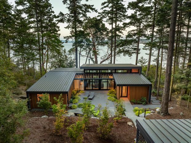 A contemporary low-profile timber-clad house nestled among mature pine trees on a wooded site, with multiple dark grey metal roof planes, extensive glazing, and a central covered outdoor courtyard or entry area. The building appears elevated on a sloping, forested site, with a body of water visible through the trees in the background.