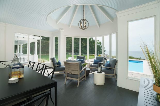 A bright enclosed sunroom or conservatory with a circular vaulted ceiling, white beadboard-style ceiling panels, exposed radial beams, and extensive windows overlooking a lawn, pool and water beyond. The room combines a dark dining table with black-framed chairs and a relaxed seating area of blue-grey armchairs around a small table. Dark flooring, white trim, and abundant glazing create an airy coastal atmosphere, with a sculptural pendant light overhead.