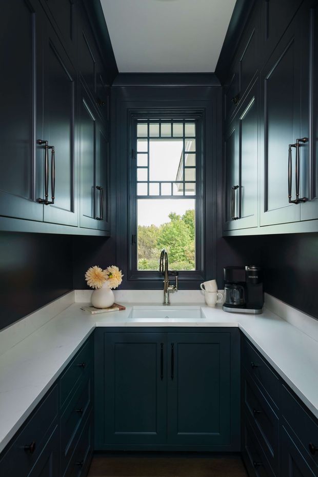 A compact galley kitchen with deep navy-blue Shaker-style cabinetry on both sides, white countertops, a centered undermount sink beneath a black-framed multi-pane window, and dark wood flooring. Copper-toned cabinet pulls, a stainless or chrome faucet, a coffee machine, and a small vase of flowers add detail. The room is symmetrical, enclosed, and softly lit, with greenery visible outside the window.