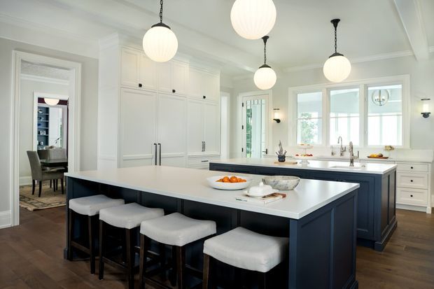 Bright, spacious classic kitchen with a large white island featuring dark navy-blue cabinetry and four upholstered stools. The room includes extensive white built-in cabinetry, a second island or peninsula with sinks and brass-toned fixtures, wide windows, glass doors, dark timber flooring, and multiple globe pendant lights. A dining area is visible through the doorway.