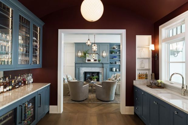 A richly styled home bar and beverage service area viewed toward an adjoining sitting room. The foreground features dark blue built-in cabinetry with glass-front display storage, a pale stone benchtop, a sink and brass-toned tap, and a collection of bottles and glassware. Deep burgundy painted walls and ceiling create a dramatic backdrop. Through the wide trimmed opening is a refined sitting room with blue built-in cabinetry, a fireplace, upholstered lounge chairs, and decorative pendant and wall lighting. White door and window trim, timber flooring, and a large white-framed window complete the scene.