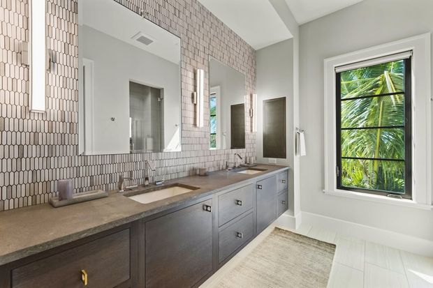 Bright, refined bathroom with a long double vanity featuring dark grey built-in cabinetry and a stone benchtop. The wall behind the vanity is clad in small pale patterned tiles, with large mirrors, twin wall lights, and two basins. White tiled flooring, a neutral rug, and a black-framed window overlooking palm foliage complete the space.