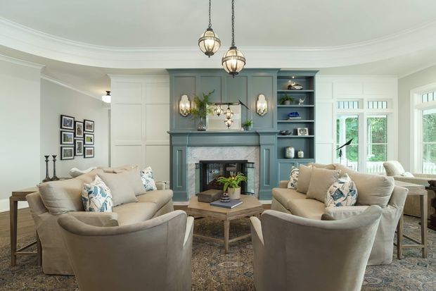 A refined traditional living room with two upholstered sofas and armchairs arranged around a low coffee table and fireplace. The room features a blue-grey painted fireplace mantel with integrated shelving, pale panelled walls, ornate ceiling cornices, patterned rug, framed artwork, and elegant pendant lighting. Tall multi-pane windows and glazed doors bring in natural light and overlook greenery.