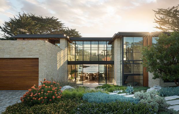 Contemporary low-profile residence with a flat roof, pale stone-clad solid walls, warm timber garage door and vertical timber accents. A dramatic double-height glazed central connection forms a transparent entry or living volume, with a visible interior beyond. The landscaped foreground includes layered drought-tolerant planting, boulders and paving, while mature trees frame the facade. Warm late-day sunlight creates a refined connection between the home and its garden.
