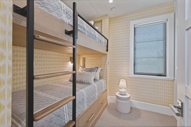 Compact bunk bedroom with a built-in timber bunk bed, black metal ladder and guard rails, neutral bedding, patterned cream wallpaper, a roller-shaded window, and a small bedside table with lamp. The room has a neat, tailored, contemporary look and carpeted flooring.