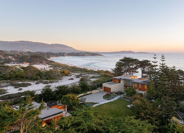 Elevated exterior view of a contemporary low-profile coastal residence set within a landscaped site, with flat roof forms, extensive glazing, timber-clad sections, pale rendered walls, and paved courtyards. The property overlooks a sandy beach, sheltered bay, and open ocean, with established trees and dense planting surrounding the buildings.