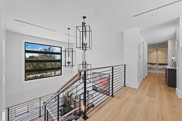 A bright, contemporary upper-level landing overlooking a stairwell. The space features pale timber flooring, white walls and ceiling, black horizontal metal stair balustrades, a timber handrail, large horizontal window, and several black geometric pendant lights. A hallway with built-in cabinetry extends toward the rear, while a glimpse of a lower level and indoor plants is visible through the railing.