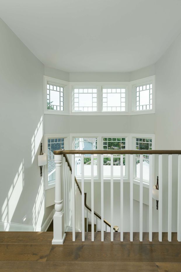 Bright, refined stair landing with a curved bay of divided-light windows, white-painted walls and trim, and a traditional staircase with turned newel post, white balusters, dark handrail, and integrated wall sconces. Warm timber flooring contrasts with the light interior, while strong daylight creates geometric window shadows on the wall.