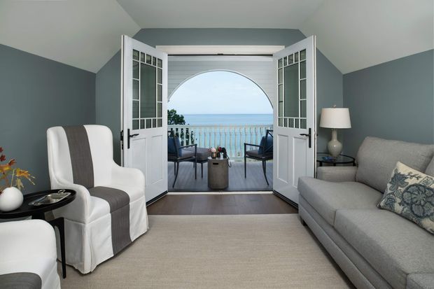 A refined coastal-style living room with deep blue-grey walls, a light neutral carpet, a grey sofa, and a white armchair with a charcoal stripe. Open French doors with divided glass panes lead to a balcony furnished with outdoor chairs and a small table, framing an ocean view through an arched opening. The room has a calm, airy atmosphere with restrained nautical detailing and soft natural light.