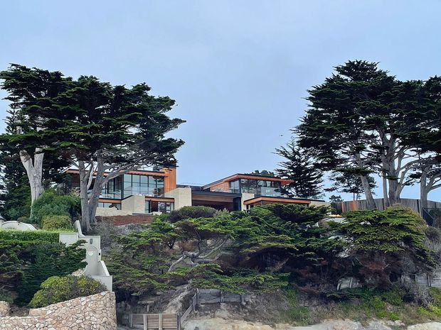 Exterior view of a multi-level contemporary home set into a landscaped, sloping coastal-style site. The building features broad glazing, warm timber-toned cladding and trim, pale masonry walls, and multiple low-pitched roof planes, partially screened by mature sculptural trees and layered garden planting.