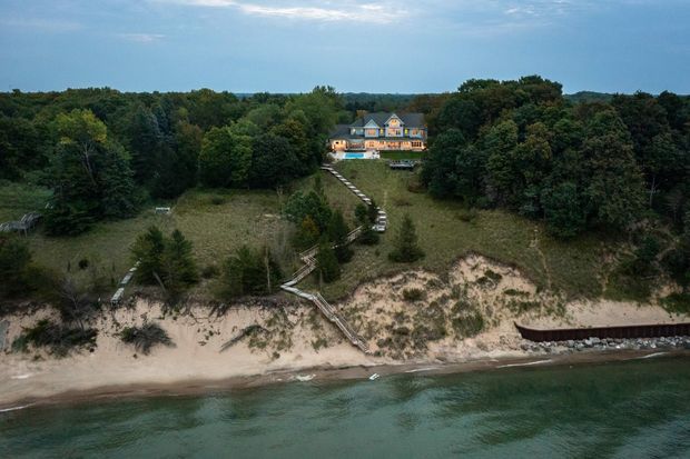 Aerial view of a large multi-storey house set within dense woodland on a sandy waterfront site. A long timber stair and walkway descend through the landscaped dune to the beach and water, with a small retaining structure along the shoreline. The home has a prominent pitched roof and multiple dormer-like roof projections, with warm lighting visible on the facade.
