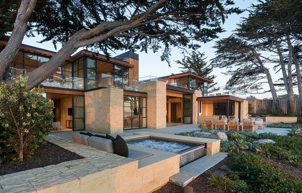 A contemporary multi-level residential house set within a landscaped coastal-looking garden, viewed from the exterior. The building features extensive floor-to-ceiling glazing, warm timber soffits and window framing, earthy masonry or stone-clad walls, projecting flat roof forms, and multiple terraces. A shallow rectangular water feature sits in the foreground beside paved paths, outdoor seating, and planted garden beds. Large mature trees partially frame the composition.