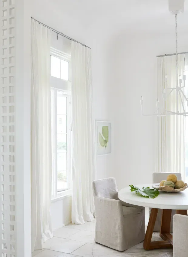 Bright, airy dining room with a predominantly white palette. The space features tall windows dressed in sheer white curtains, a white round dining table with upholstered slipcovered chairs, a timber pedestal base, a contemporary linear chandelier, white tiled flooring, and a decorative breeze-block screen along the left wall. Minimal botanical artwork and a bowl of fruit add restrained natural accents.