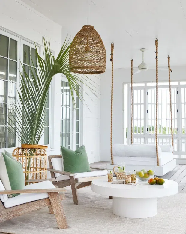 Bright, airy coastal-style living room with white painted walls, large multi-pane windows and doors, pale timber flooring, rattan lounge chairs, a woven pendant light, green cushions, and a white suspended swing sofa. A round white coffee table with fruit and drinks anchors the seating area, while a tall palm adds a tropical accent.