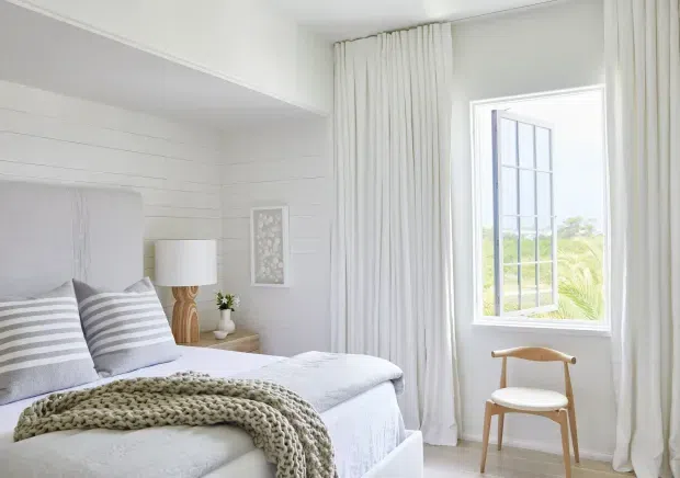 Bright, serene bedroom with white horizontal wall panelling, a white upholstered bed, layered grey-and-white striped pillows, and a chunky knitted throw. Full-height white curtains frame an open multi-pane window, while a simple wooden chair, bedside table, sculptural lamp, and small decor create a soft coastal-inspired setting.