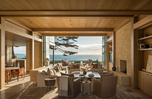 A warm, refined living room with a large open glazed wall framing a coastal ocean view. The room features extensive natural timber lining on the ceiling and structural beams, a substantial textured stone fireplace wall, built-in timber shelving and cabinetry, and a layered seating arrangement around a central coffee table. An adjoining dining area is visible to the left through an open connection, while outdoor seating appears beyond the glazing.