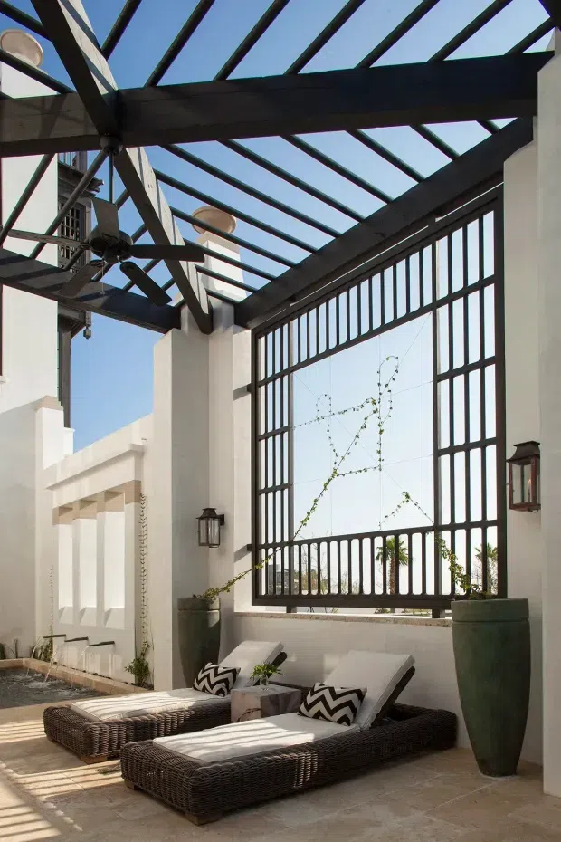An airy outdoor courtyard or covered terrace with white rendered walls, a dark exposed-beam pergola and glazed roof structure, and a large black metal grid window or screen. Two woven chaise lounges with patterned cushions sit on a pale tiled floor, framed by tall planters and wall-mounted lanterns. The space has strong geometric shadows, abundant daylight, and a refined Mediterranean-inspired feel.