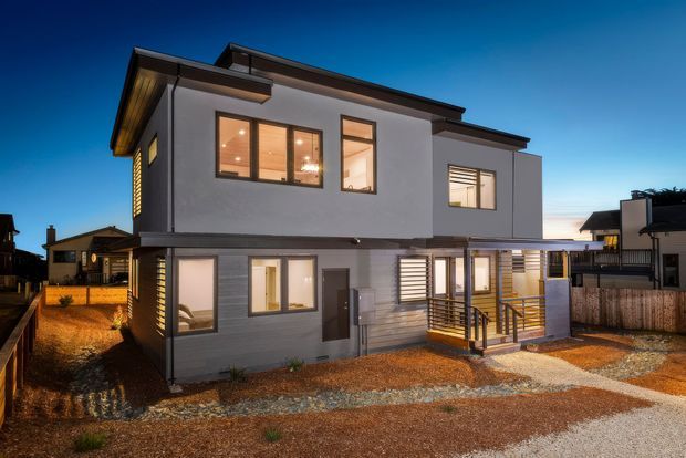 Twilight exterior photograph of a contemporary two-storey residential house. The home has a clean geometric form, flat and low-pitched roof sections, smooth light-grey rendered upper walls, darker horizontal lower cladding, large rectangular windows, and a timber-toned entrance deck with metal balustrading. Gravel landscaping and neighbouring homes surround the property.
