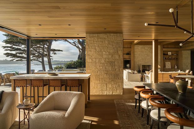 A warm, open-plan coastal living and dining space with expansive glazing overlooking the ocean. The room features a timber-lined ceiling, dark timber flooring, a textured stone fireplace wall, a kitchen island with bar seating, a dining table, sculptural upholstered lounge chairs, and contemporary pendant lighting. Natural light and views connect the interior to the landscaped coastal setting.
