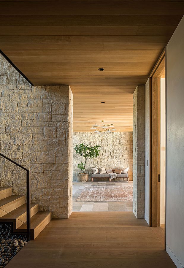A warm, refined residential interior viewed from a timber-floored hallway or entry passage. Rough-cut natural stone walls frame an opening toward a relaxed living room with a sofa, rug, and indoor plant. The space features extensive timber slat ceilings, recessed downlights, timber flooring, a black metal stair balustrade, and a partially visible staircase descending beside a pebble-filled feature area. Natural light enters through glazing on the right, creating a calm, earthy material palette.