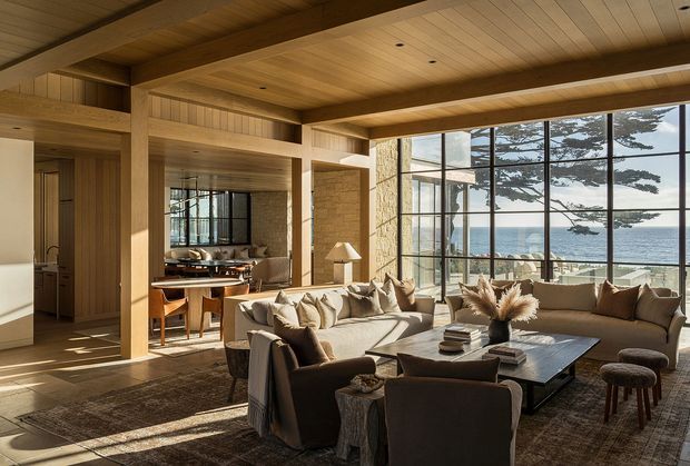 Warm, sophisticated open-plan living and dining area with extensive floor-to-ceiling glazing overlooking the ocean. The room features a heavily timber-lined ceiling and structural beams, vertical timber wall panelling, natural stone walls, dark framed glazing, a large neutral sectional sofa, dining table, lounge chairs, and layered rugs. Strong natural light and the coastal outlook create an inviting, refined atmosphere.