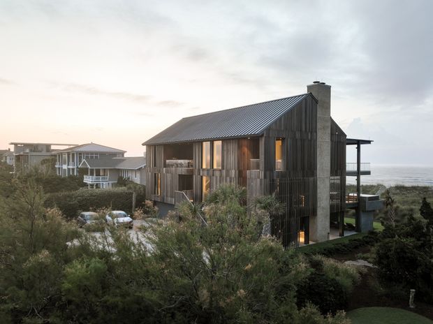 Exterior view of a contemporary coastal house set among dense dune-like landscaping, with dark vertical timber cladding, a steep corrugated metal gable roof, tall concrete chimney, and large glazed openings. The ocean is visible beyond the property, with neighbouring coastal homes nearby.