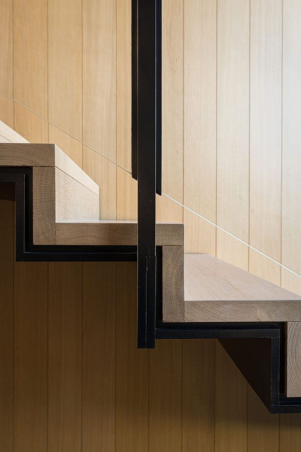 Close-up architectural photograph of a modern staircase with pale timber-clad walls and stair treads, contrasted by exposed black-painted steel stringers and supports. The stair structure has a crisp, floating or zigzag profile with minimal detailing and strong linear geometry.