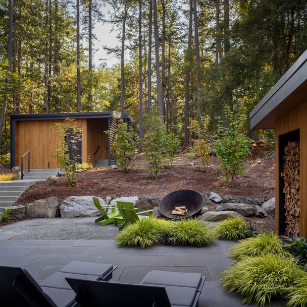 A landscaped woodland outdoor living area with dark paved terrace, low black lounge furniture, green Adirondack chairs, a round fire bowl, ornamental grasses, stone retaining walls, and two compact timber-clad structures nestled among tall trees. One structure has an open wood-storage alcove beneath a sloping roof.