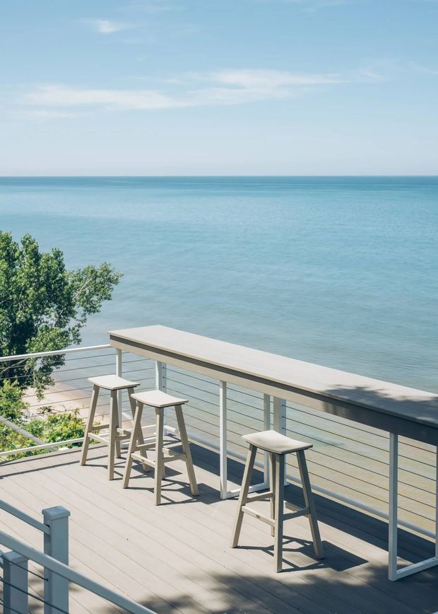 A bright, elevated outdoor deck overlooking a calm blue sea. The deck has pale timber-look flooring, streamlined horizontal metal railings, and a long built-in bar-height counter with several stools. Trees and a small glimpse of shoreline appear below, creating an airy coastal entertaining setting.