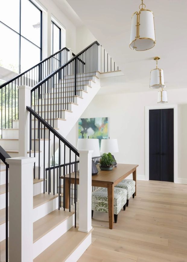 Bright, refined open-plan entry and stair hall with a prominent sculptural staircase featuring pale timber treads, white stringers and posts, and slim black metal balustrades. Full-height glazing brings in abundant natural light. A timber dining table with upholstered patterned chairs sits beneath a series of elegant white-and-gold pendant lights, with a dark internal door and contemporary artwork adding contrast.