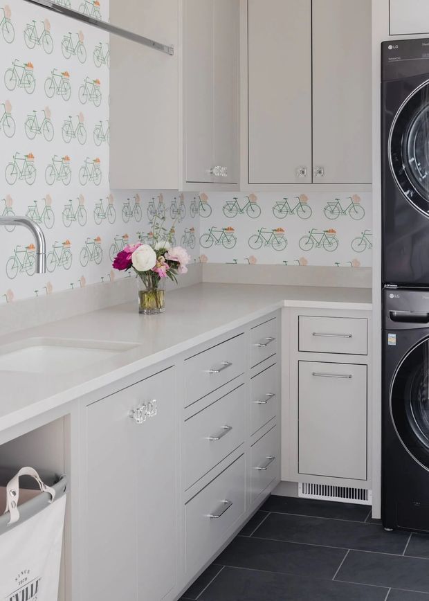 Bright, compact laundry room with extensive pale cabinetry, a light solid-surface L-shaped benchtop, integrated sink, patterned bicycle-print splashback, chrome hardware, dark floor tiles, and a stacked black front-loading washer and dryer.