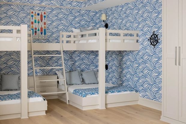 A bright children's bedroom with two white bunk-bed structures, integrated ladders and under-bed drawers, pale timber flooring, and white built-in wardrobe cabinetry. The room features a bold blue-and-white wave-pattern wallpaper and coordinated blue patterned bedding, with nautical wall artwork and a compass decoration adding a maritime theme.
