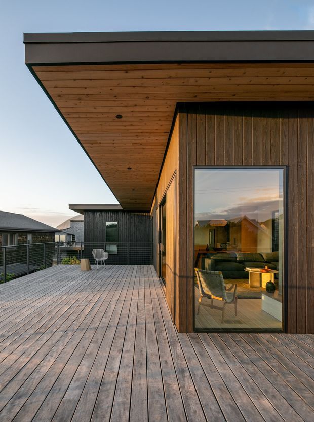 Exterior architectural photograph of a contemporary timber-clad dwelling with a broad flat roof overhang and a large timber deck terrace. The image focuses on the deck and facade, with vertical dark timber wall cladding, warm timber soffit lining, dark metal roof edging, and full-height glazed doors revealing a living room inside. A neighbouring dwelling is visible in the background.