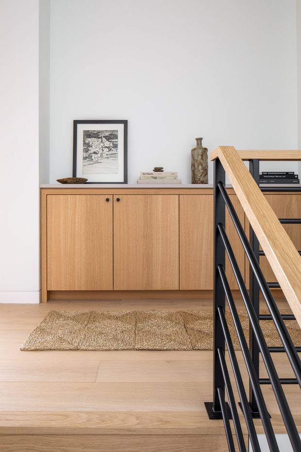 A refined interior landing or hallway beside a staircase, featuring a low, light timber built-in cabinet with flat-panel doors and dark knobs against a white wall. The cabinet is styled with framed artwork, books, a ceramic vase and a shallow bowl. Pale timber flooring and a natural woven rug create a warm, understated setting, while the foreground staircase has a black metal balustrade and a light timber handrail.