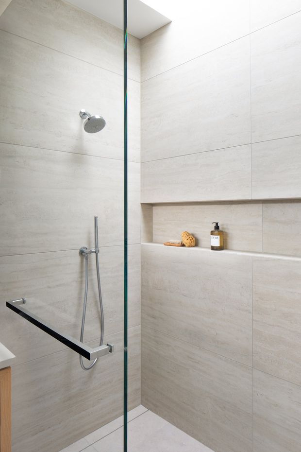 Minimalist walk-in shower with large pale stone-look wall tiles, a frameless glass screen, chrome rain shower and handheld shower, and a recessed shelf with toiletries and bath accessories. The space is bright, clean, and understated, with a concealed ceiling recess above the shower.