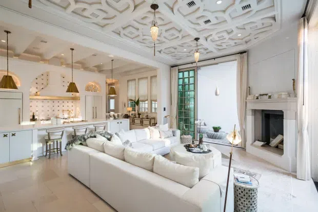 Bright, luxurious open-plan living space combining a white kitchen, dining area and large living room. The room features an ornate geometric coffered ceiling, tall windows with layered curtains, a decorative fireplace with white mantel, pendant lighting, pale flooring and a large white sectional sofa. The kitchen includes a central island, pale cabinetry and a softly lit display niche.