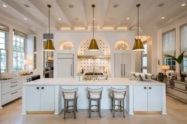 Bright, spacious luxury kitchen centered on a large white island with seating for three. The room features white perimeter cabinetry, integrated appliances, a decorative tiled or stone backsplash with arched niches, brass/gold pendant lights and hardware, pale timber flooring, detailed coffered ceiling treatment, and large multi-pane windows. The styling is refined and classic with warm metallic accents.