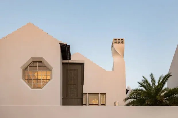 Exterior view of a pale stucco residence with a stepped gable-like roofline, dark timber entrance door, distinctive octagonal multi-pane window, small lower window, and a sculptural chimney. A palm tree is visible behind the boundary wall against a clear blue sky.