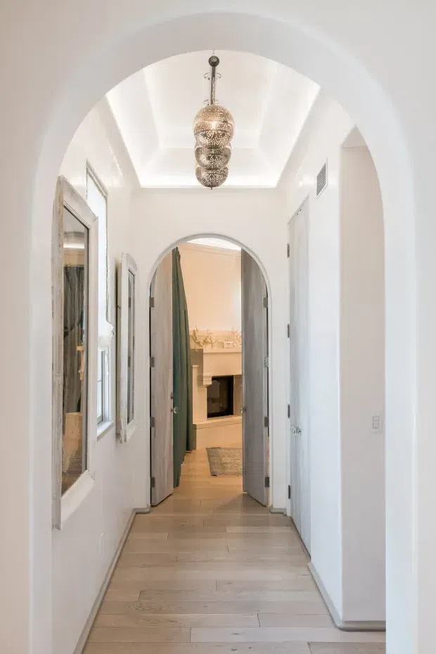 Bright, narrow residential hallway viewed through a large rounded archway. The corridor has white painted walls, arched door openings, pale timber-look flooring, white doors and trim, mirrored wall panels on the left, and a decorative stacked pendant light hanging from a recessed tray ceiling. An adjoining room with a fireplace is visible at the end of the hall.