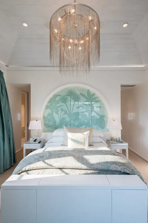 A bright, refined bedroom viewed head-on, featuring a large bed with pale blue and white patterned bedding, matching bedside tables and lamps, a curved decorative feature wall behind the bed with botanical imagery, teal curtains, pale timber flooring, and a dramatic tiered metallic pendant chandelier. The room has a soft coastal-inspired palette and abundant natural light.