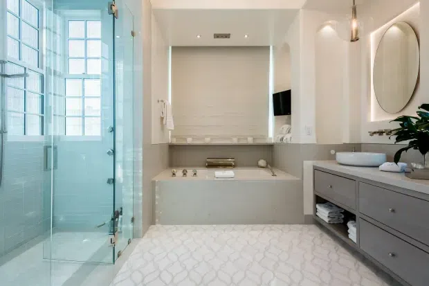 Bright, refined contemporary bathroom featuring a glass-enclosed shower at left, a freestanding-style bathtub set into a tiled alcove, and a wide grey double vanity with integrated storage at right. The room has pale textured wall tiles, light stone-look surfaces, arched recessed niches, oval mirrors with backlighting, chrome fixtures, and a patterned light floor. Large frosted or textured windows bring in abundant natural light.