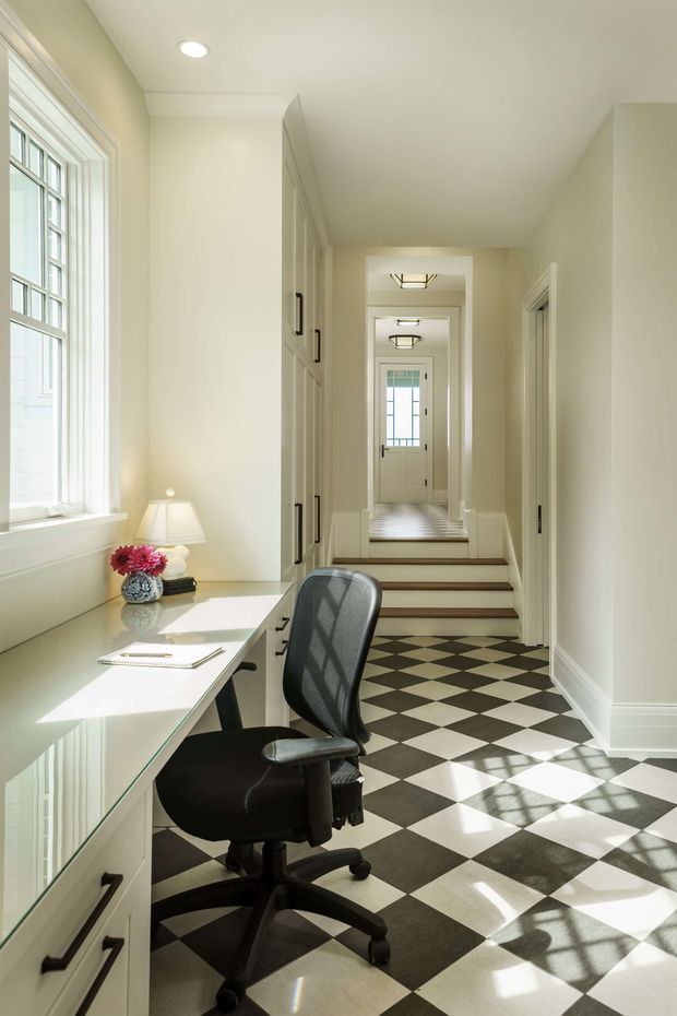 A bright, narrow home office positioned along a long hallway. A white built-in desk runs beneath a large multi-pane window, with a black ergonomic office chair, small table lamp and flowers. The hallway continues beyond several steps and doorways, with distinctive black-and-white checkerboard flooring, pale cream walls, white trim and glazed interior doors. Natural sunlight creates strong geometric shadows across the floor.