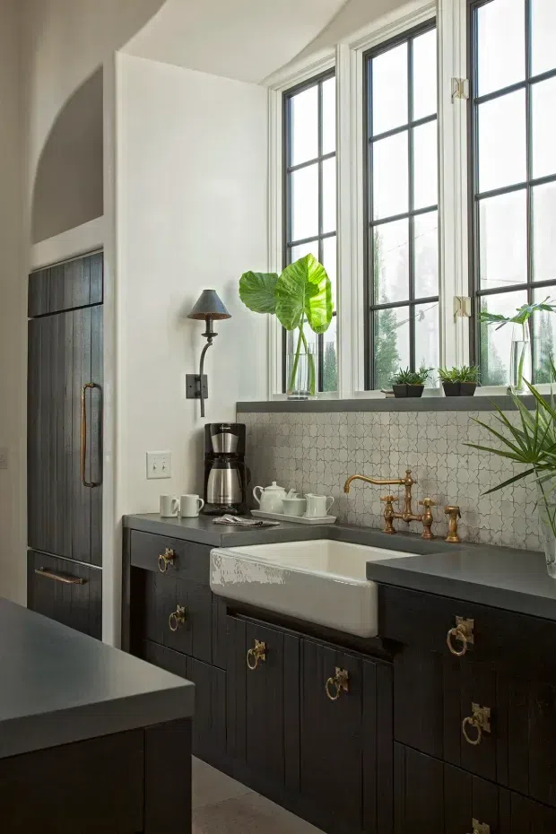 A refined kitchen coffee and wash-up station with dark vertically grooved cabinetry, a pale grey countertop, a large white farmhouse apron-front sink, and an ornate aged-brass gooseneck tap. The backsplash features small light patterned tiles, while tall black-framed multi-pane windows bring in abundant daylight. A coffee maker, crockery, potted plants, and a wall-mounted sconce add functional and decorative detail. The space has a traditional, tailored character with a muted, high-contrast palette.